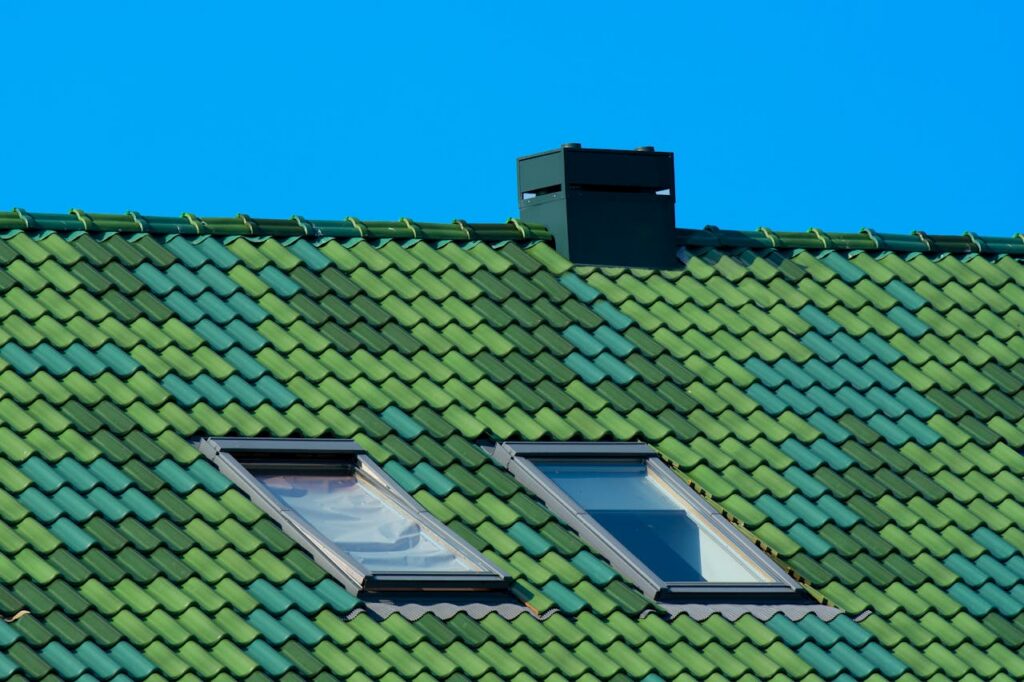 A modern architectural roof with green tiles, skylights, and chimney against a blue sky.